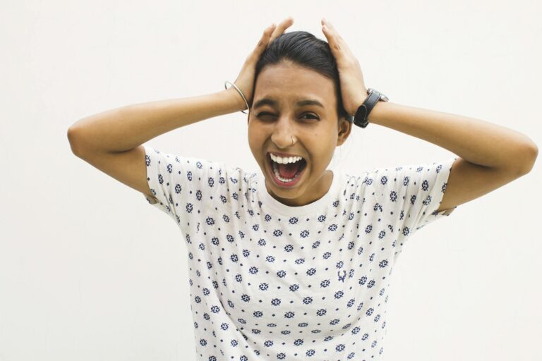 A young woman in a casual shirt screams with hands on her head against a white background.